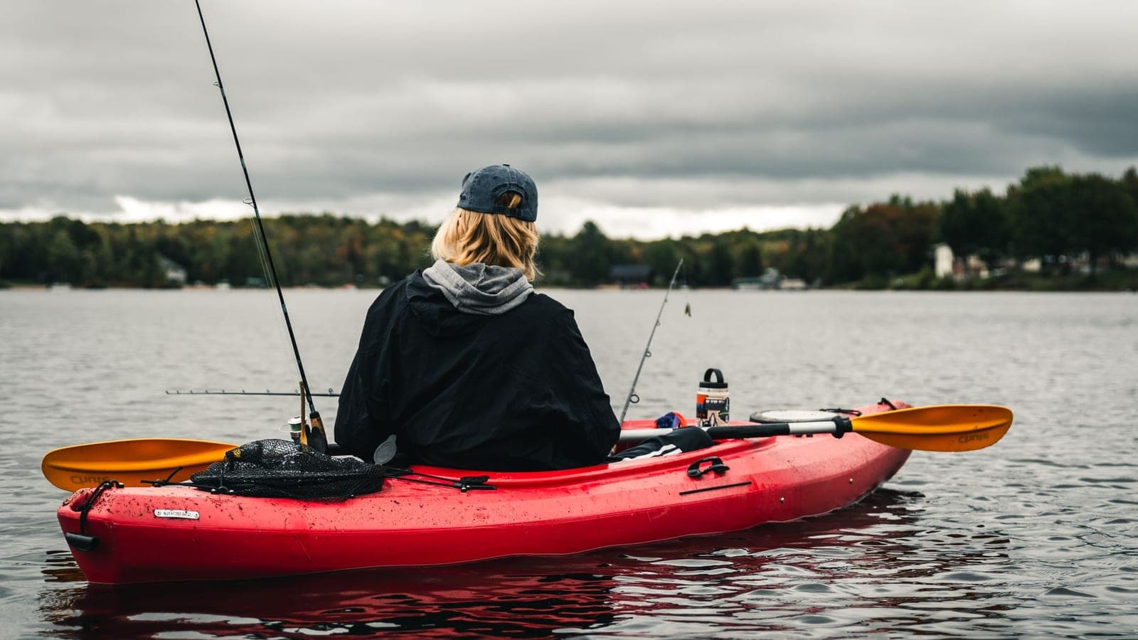 An angler seated in a red kayak on a calm lake under cloud