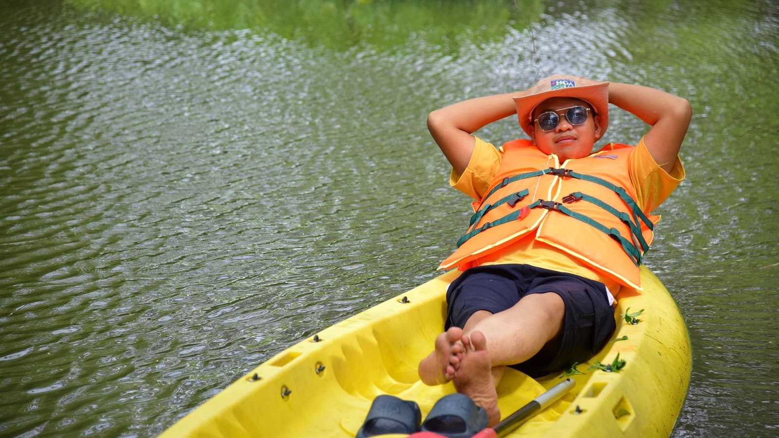 A paddler in an orange life jacket sitting in a yellow kayak on a calm lake