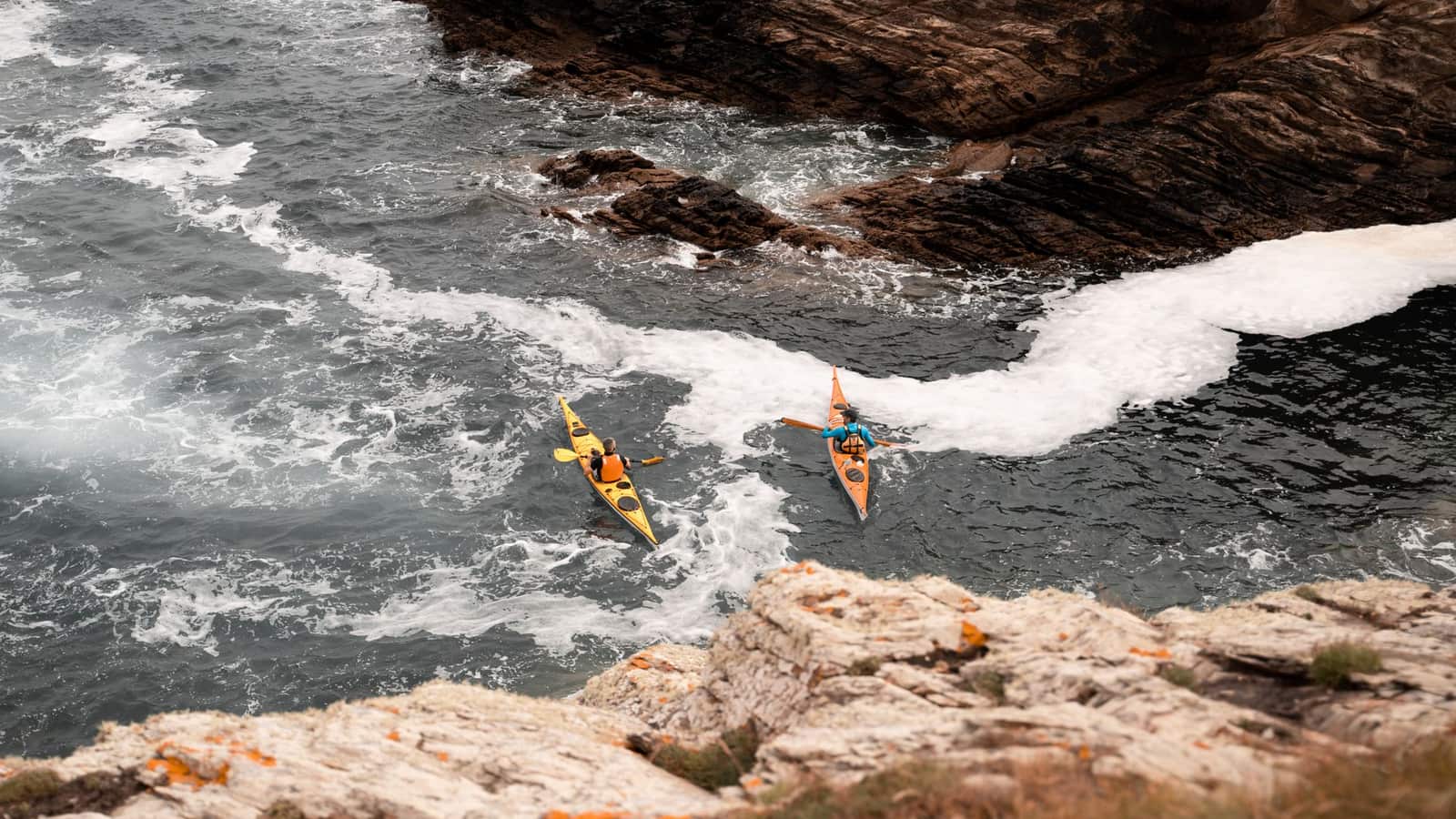 Two kayakers paddling through small waves near a rocky coastline