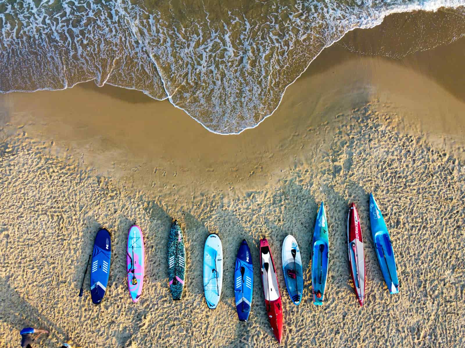 A hard epoxy paddle board lying on sand next to the water
